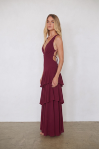 A woman modeling a sleeveless, deep burgundy maxi dress with a tiered ruffle skirt and side cutouts, standing against a plain white wall.