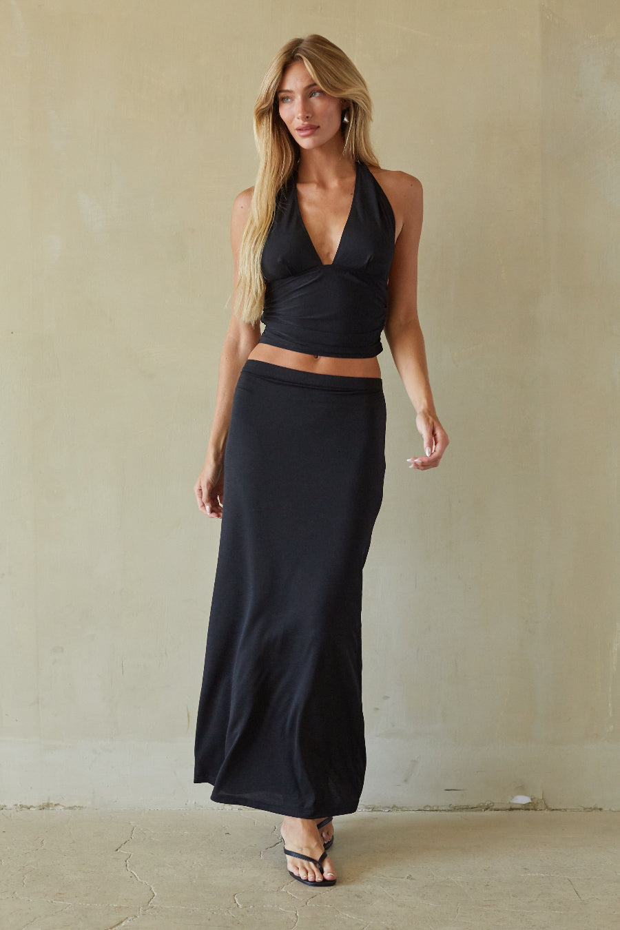 A woman modeling a black two-piece outfit consisting of a halter wrap-style crop top and a matching long maxi skirt, standing against a textured beige wall.