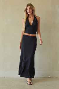 A woman modeling a black two-piece outfit consisting of a halter wrap-style crop top and a matching long maxi skirt, standing against a textured beige wall.