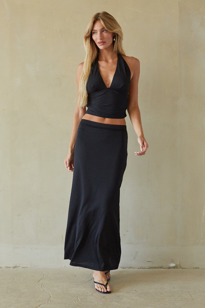 A woman modeling a black two-piece outfit consisting of a halter wrap-style crop top and a matching long maxi skirt, standing against a textured beige wall.
