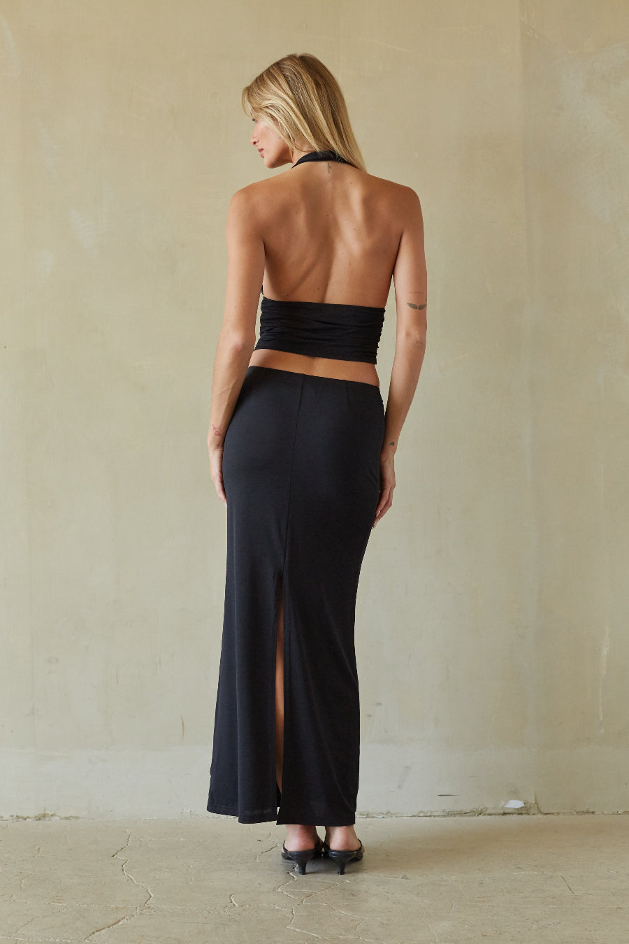 A woman stands with her back to the camera wearing a black two-piece outfit consisting of a halter-style bandeau top and a matching high-slit maxi skirt, set against a neutral beige textured wall.