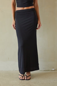 A woman wearing a long black wrap-style maxi skirt with a matching black crop top, standing against a textured beige wall.