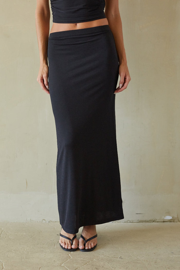 A woman wearing a long black wrap-style maxi skirt with a matching black crop top, standing against a textured beige wall.