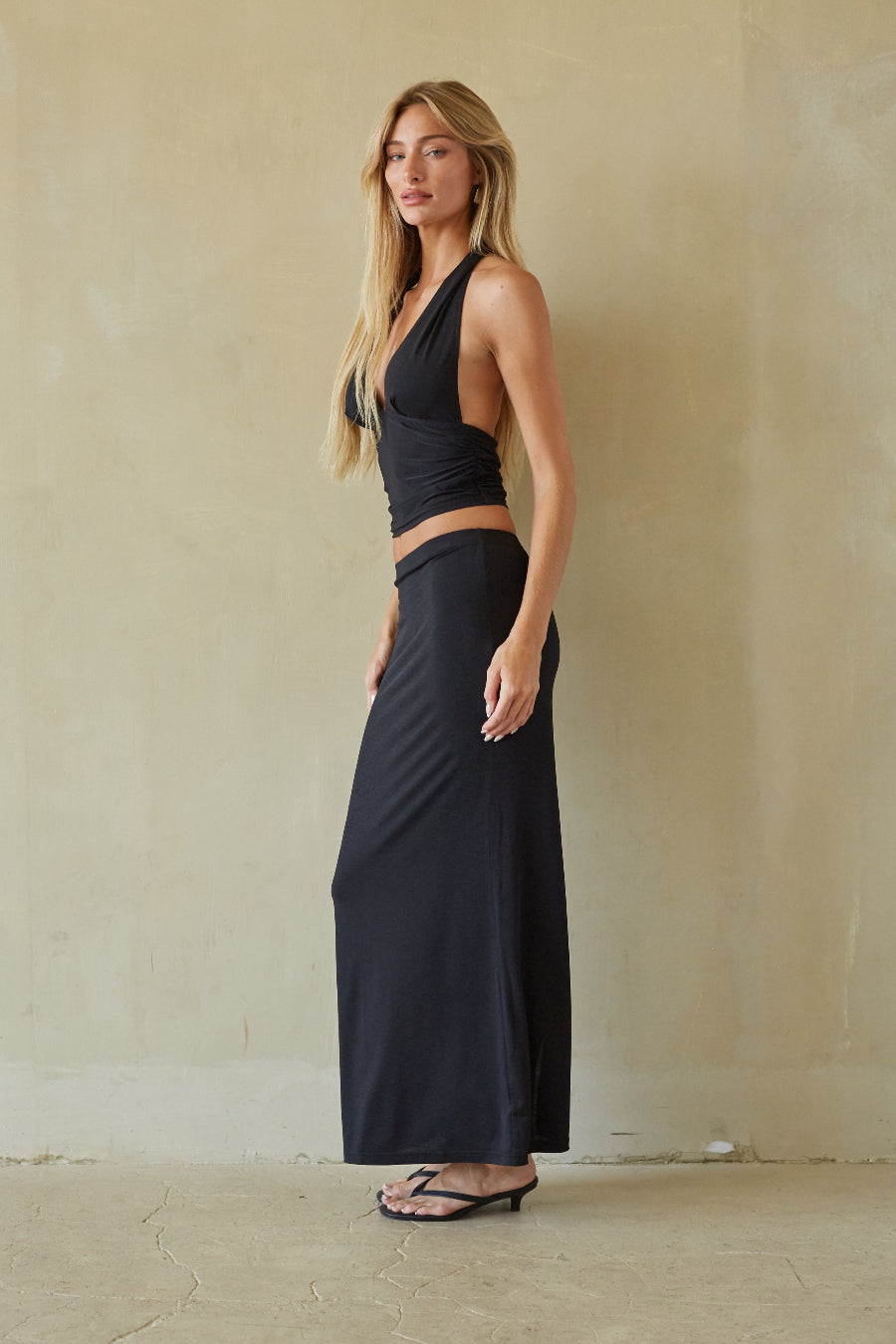 A woman modeling a black two-piece set consisting of a halter-style wrap crop top and a matching high-waisted maxi skirt, standing against a textured beige plaster wall.