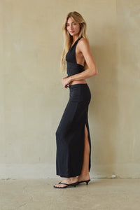 A woman modeling a black halter-neck maxi dress with a side slit, standing against a textured beige plaster wall.