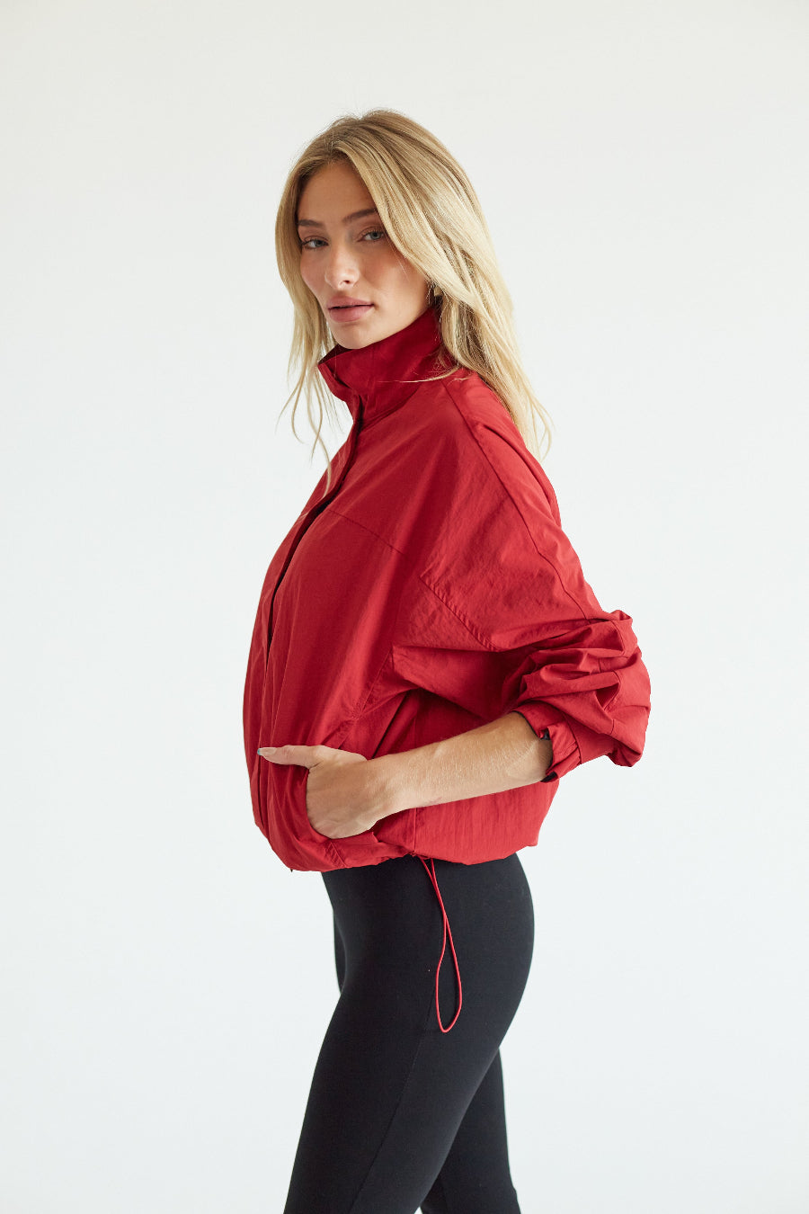 A blonde woman wearing a bright red high-neck windbreaker jacket with voluminous sleeves and black leggings, standing against a plain white studio background.