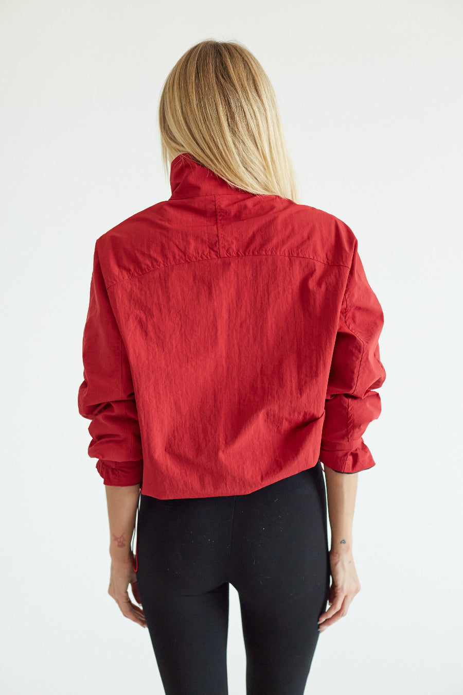 A woman viewed from behind wearing a cropped red long-sleeve shirt with a high collar and black leggings, set against a plain white studio background.