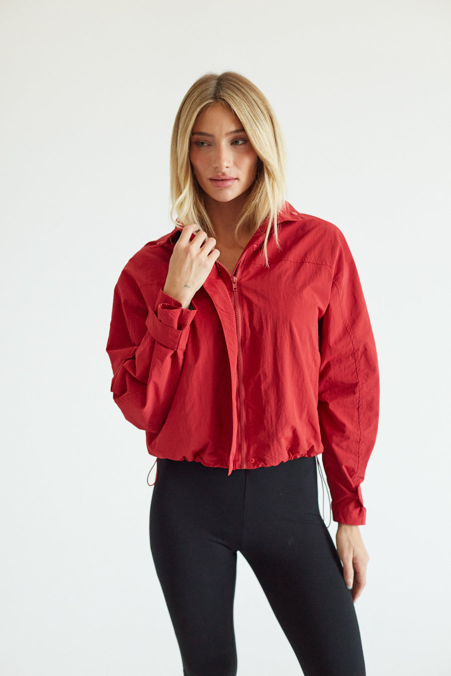 A woman modeling a lightweight red zip-up jacket with long sleeves, paired with black leggings, against a plain white studio background.
