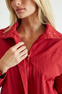 Close-up of a woman wearing a bright red lightweight zip-front jacket, with her hand adjusting the zipper. The photo is cropped from the lower face to the torso against a clean light background.