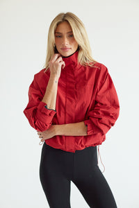 A blonde model wearing a cropped red lightweight jacket with a high collar and black leggings, posing against a clean white studio background.