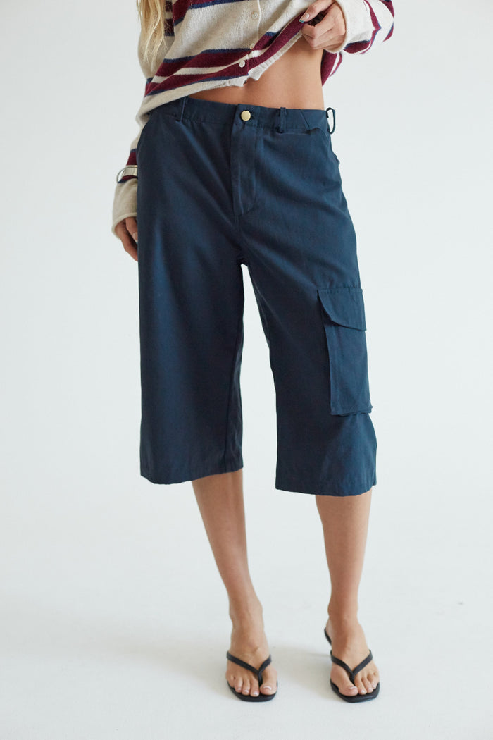 A model wearing dark navy blue cropped cargo pants with a striped knit top and black flip-flop sandals, standing against a plain white studio background.