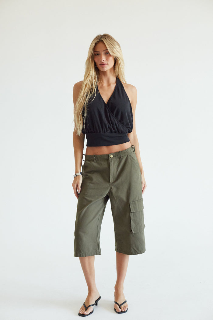 A blonde model stands against a plain white studio background wearing an olive green cropped cargo pants with side flap pockets, a black halter crop top, and black flip-flop sandals.
