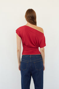 A woman viewed from the back wearing a bright red one-shoulder top and dark blue jeans against a plain white background.