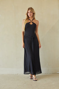 A woman modeling a black halter-neck maxi dress with a keyhole cutout at the chest and a side tie detail, standing against a textured beige wall.