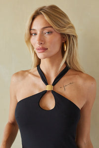 A blonde woman wearing a black halter-neck top with a gold-toned connector at the collar, photographed against a warm beige wall.