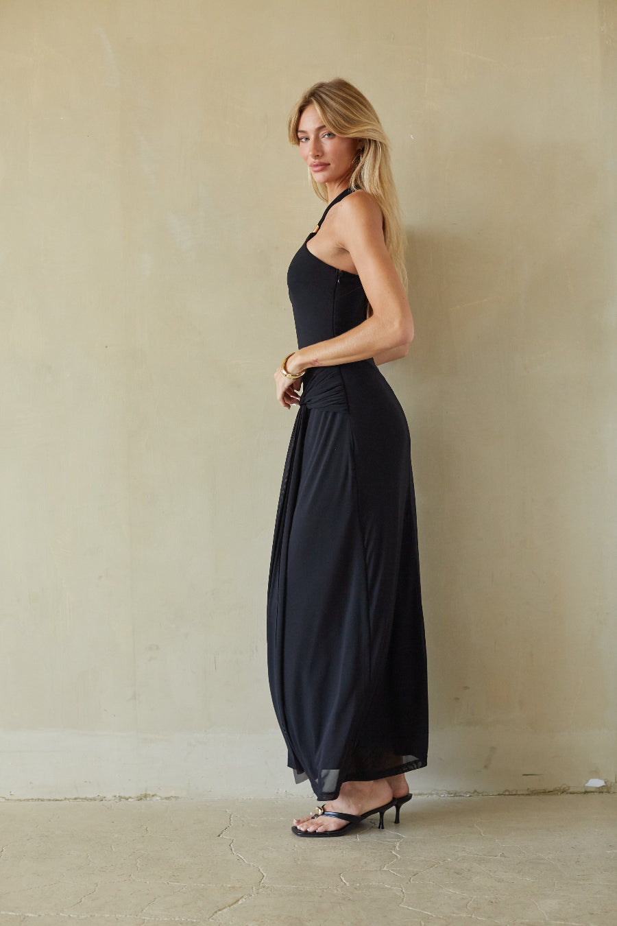 A woman modeling a black halter-neck maxi dress with a gathered waist, standing against a textured beige plaster wall.