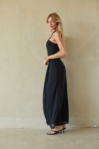 A woman modeling a black halter-neck maxi dress with a gathered waist, standing against a textured beige plaster wall.