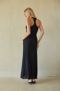 A woman modeling a black razorback maxi dress, standing against a textured beige wall.