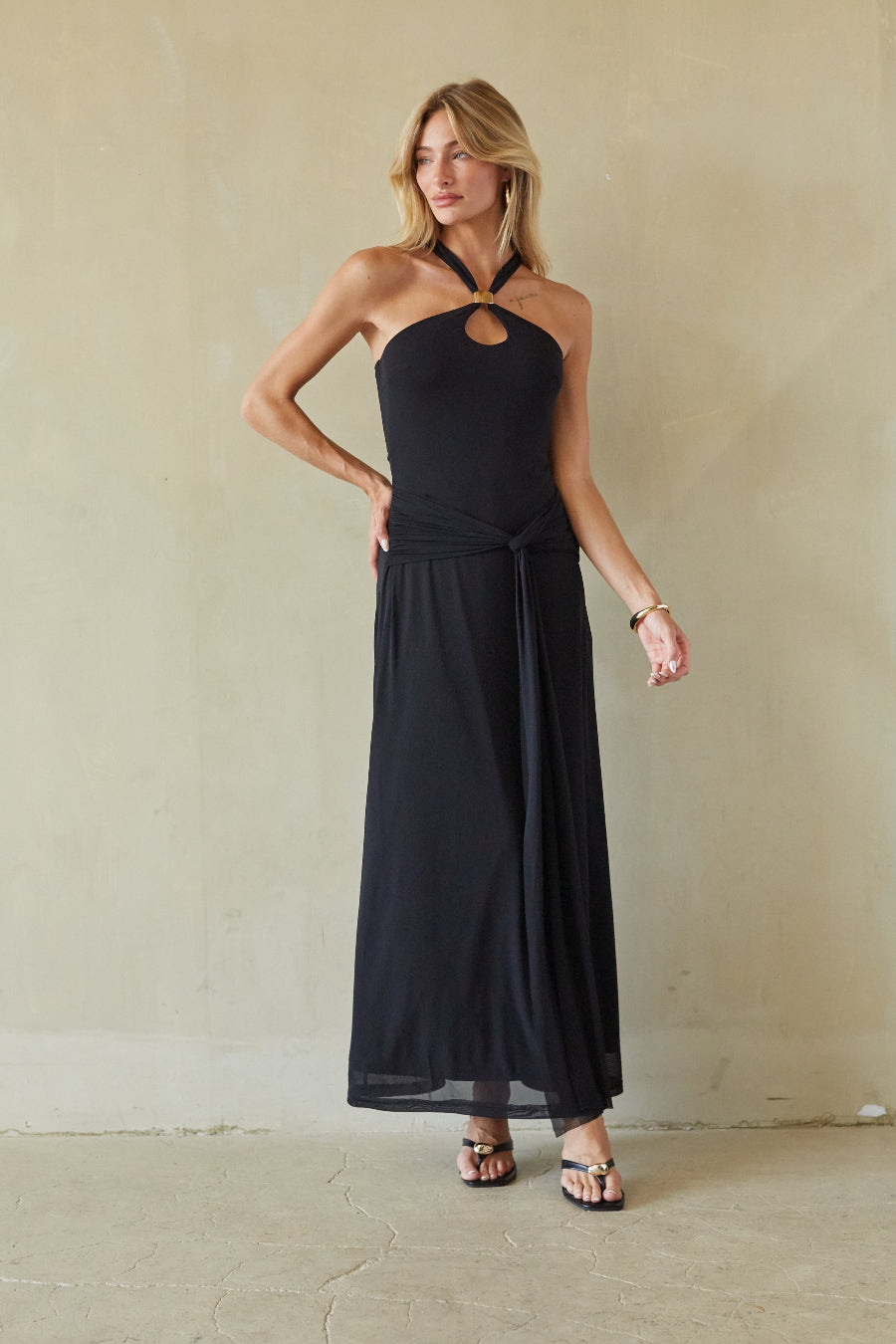 black-image | A woman modeling a black halter-neck maxi dress with a keyhole cutout at the chest and a side tie detail, standing against a textured beige wall.