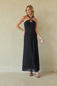 black-image | A woman modeling a black halter-neck maxi dress with a keyhole cutout at the chest and a side tie detail, standing against a textured beige wall.