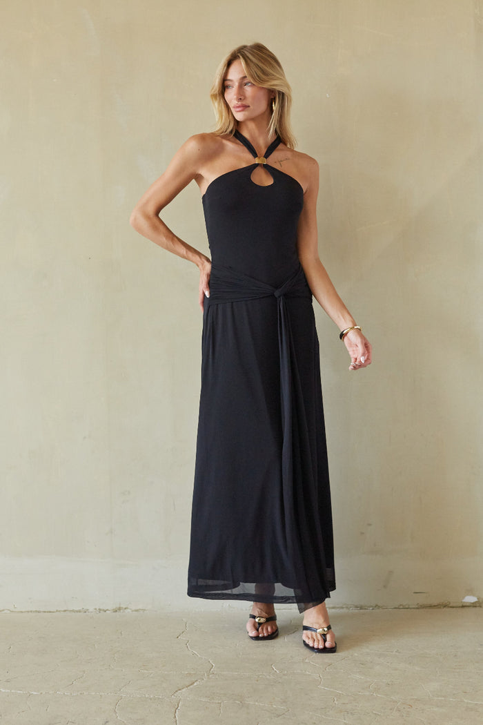 black-image | A woman modeling a black halter-neck maxi dress with a keyhole cutout at the chest and a side tie detail, standing against a textured beige wall.