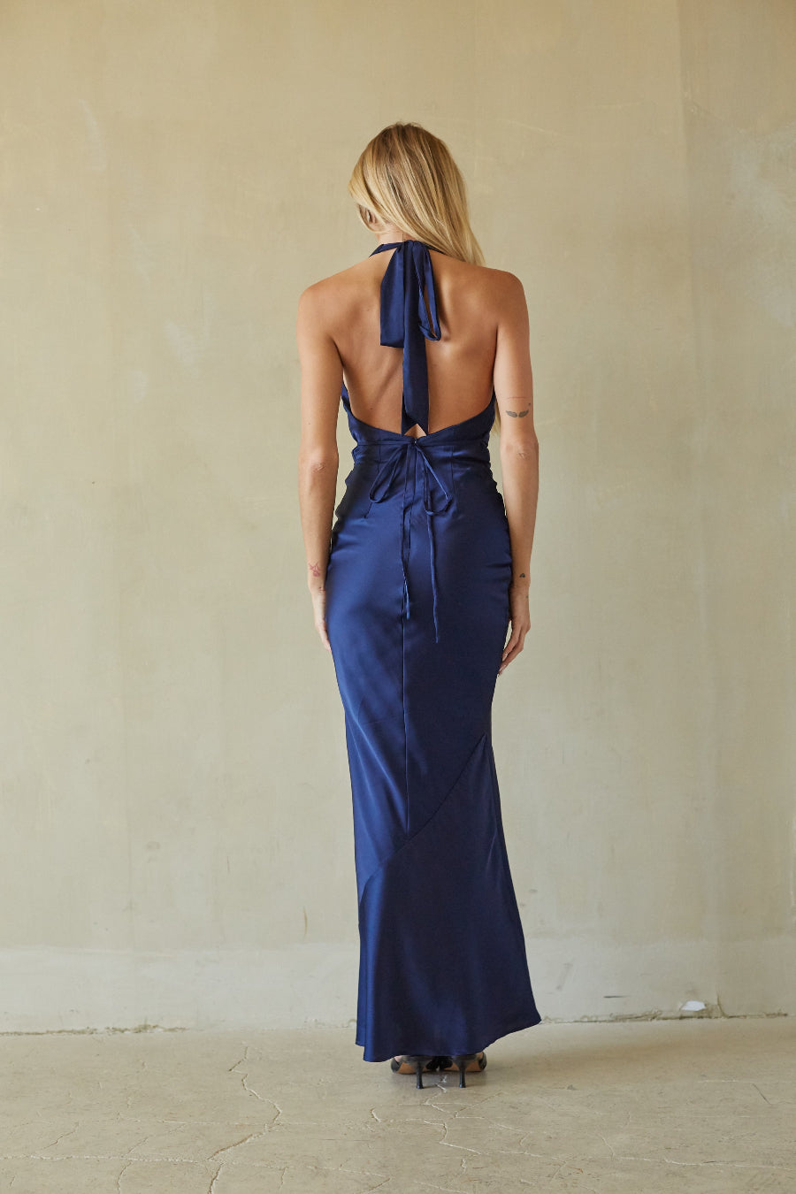 A woman viewed from behind wearing a navy blue satin halter maxi dress with an open back and tie detail, standing against a textured beige wall.
