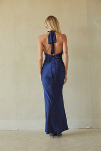 A woman viewed from behind wearing a navy blue satin halter maxi dress with an open back and tie detail, standing against a textured beige wall.