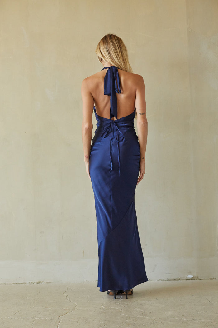 A woman viewed from behind wearing a navy blue satin halter maxi dress with an open back and tie detail, standing against a textured beige wall.
