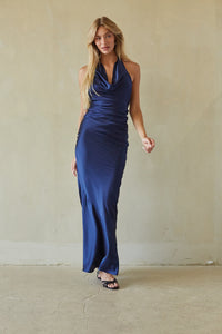 blue-image | A woman modeling a sleeveless, cowl-neck maxi dress in a rich royal blue satin, standing against a textured beige plaster wall.