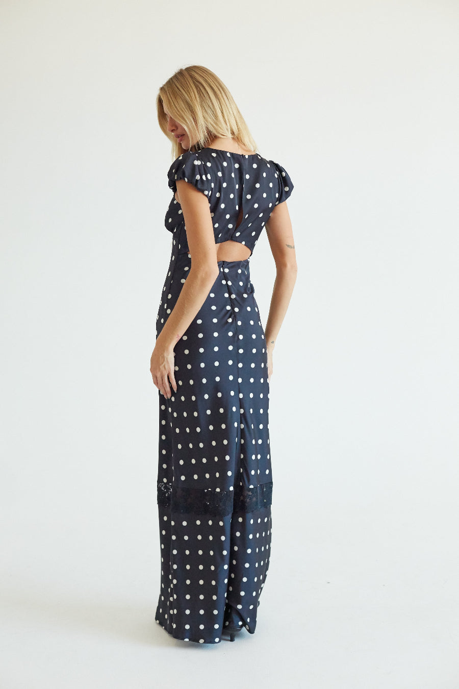 A woman wearing a navy blue maxi dress with white polka dots, shown from the back against a plain white studio background.