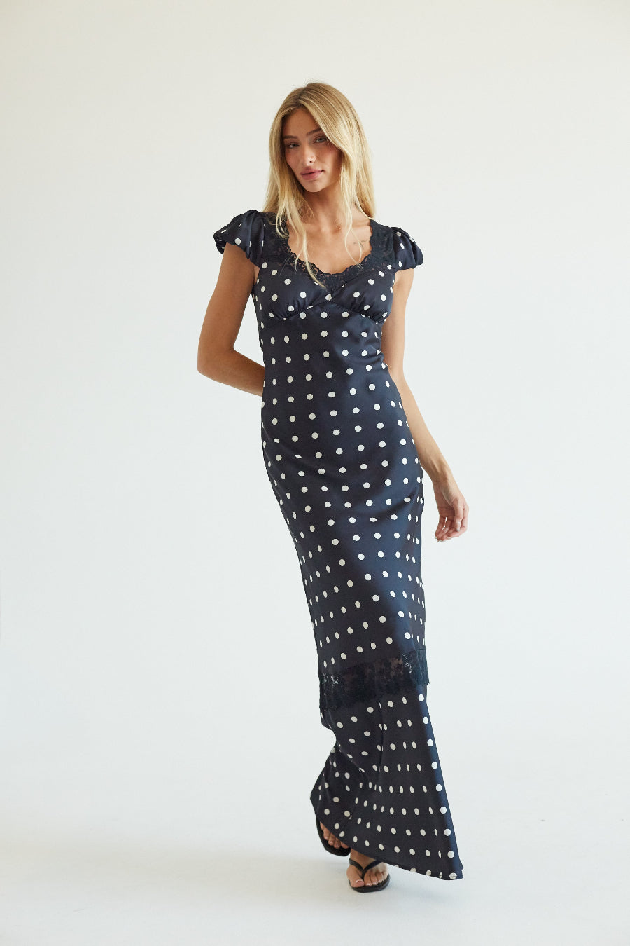 A woman modeling a navy blue maxi dress with white polka dots, ruffled cap sleeves, and a V-neckline, set against a plain white studio background.