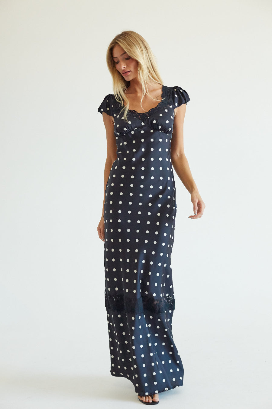 A woman modeling a navy blue maxi dress with a white polka dot pattern, featuring short puff sleeves and a ruffled neckline, set against a plain white studio background.