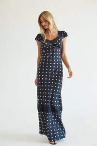 A woman modeling a navy blue maxi dress with a white polka dot pattern, featuring short puff sleeves and a ruffled neckline, set against a plain white studio background.