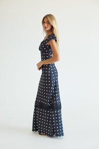 A woman modeling a navy blue maxi dress with a white polka dot pattern, standing against a plain white studio background.
