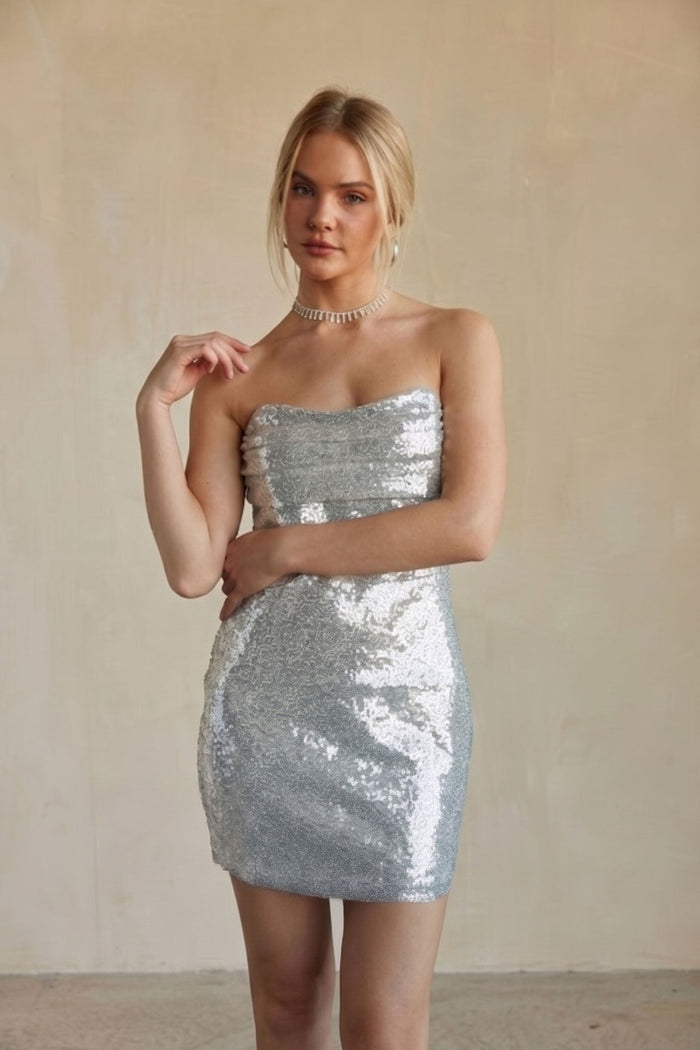 A woman modeling a strapless silver sequin mini dress against a neutral beige textured wall.