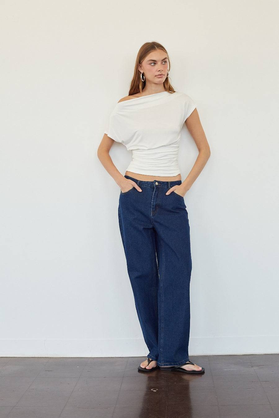 A woman stands against a plain white wall wearing a white off-the-shoulder wrap top and dark blue wide-leg jeans, with her hands in her pockets.