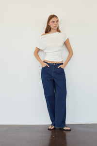 A woman stands against a plain white wall wearing a white off-the-shoulder wrap top and dark blue wide-leg jeans, with her hands in her pockets.