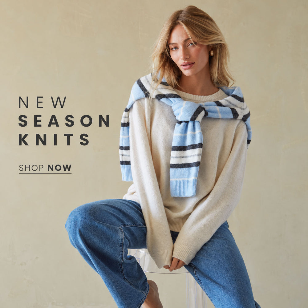 A woman seated against a warm beige wall wearing a cream knit sweater, blue jeans, and a light blue striped knit scarf. Text on the left reads 'NEW SEASON KNITS' and 'SHOP NOW'.