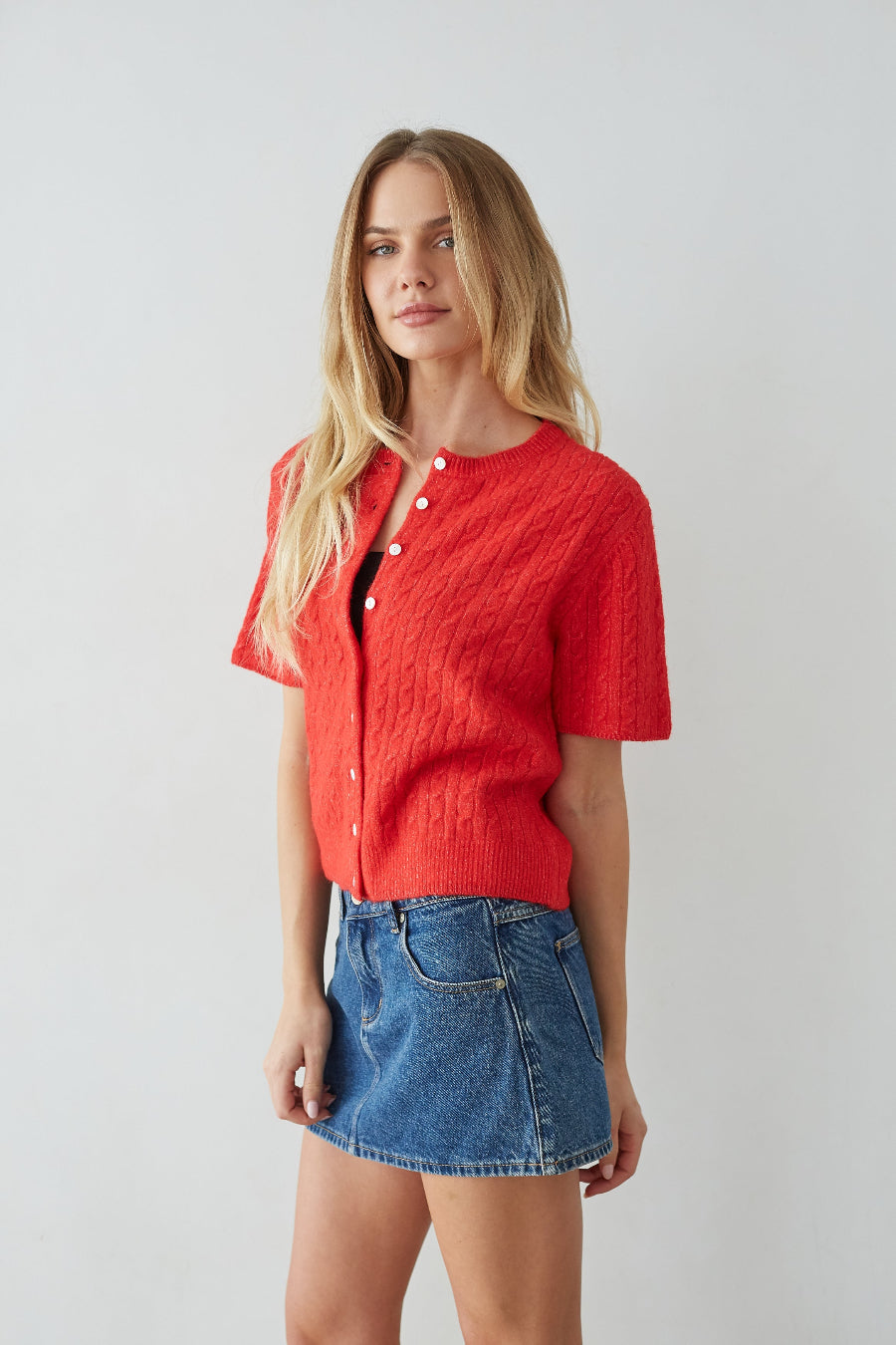 Woman wearing a red knitted cardigan over a black top with a blue denim skirt on a light gray background