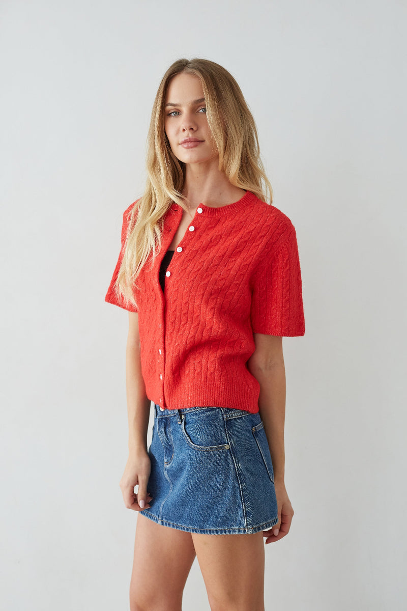 Woman wearing a red knitted cardigan over a black top with a blue denim skirt on a light gray background