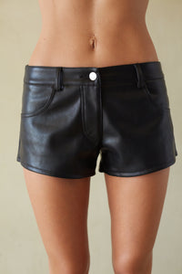 Black faux leather shorts with a silver button closure, modeled on a person against a neutral beige background.