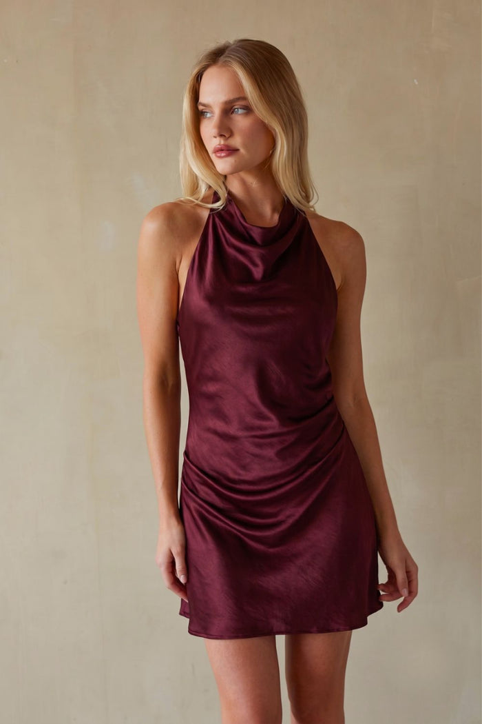 red-image | A woman modeling a sleeveless, high-neck mini dress made of draped satin-like fabric in a deep burgundy color, set against a neutral beige textured wall.