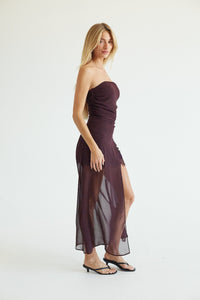 A woman modeling a strapless, dark plum-brown midi dress with a ruched bodice, sheer draped skirt overlay, and side slit, paired with black flat sandals against a plain white studio background.