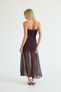 A woman viewed from behind wearing a strapless dark plum midi dress with a sheer overlay skirt, paired with black heels, against a plain white studio background.