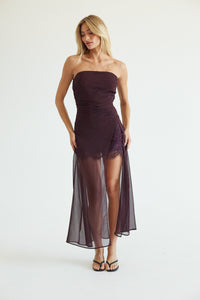 A woman modeling a strapless dark plum midi dress with a sheer mesh overlay skirt, lace bodice, and side slit, photographed against a plain white studio background.