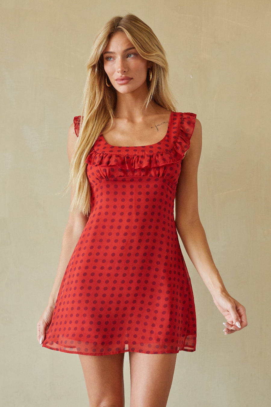 A woman modeling a red sleeveless mini dress with a black polka dot pattern and ruffled neckline, set against a neutral beige background.