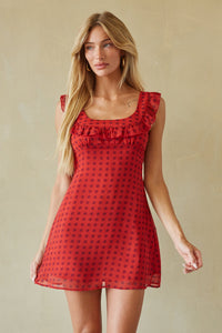 A woman modeling a red sleeveless mini dress with a black polka dot pattern and ruffled neckline, set against a neutral beige background.