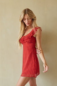 A woman modeling a red one-shoulder ruffle dress with a black polka dot pattern, posed against a neutral beige wall.