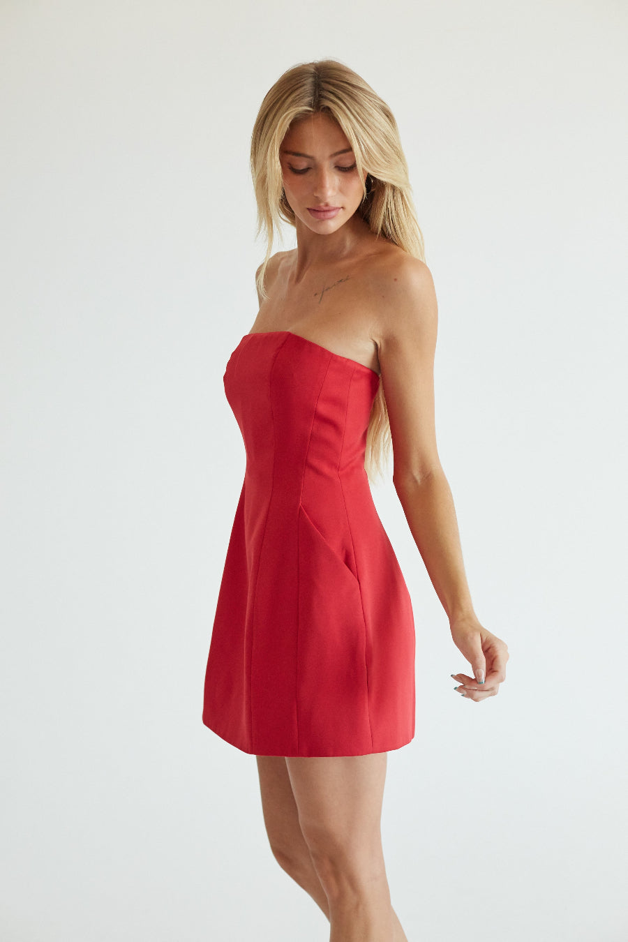 Blonde model in strapless red mini dress showing side angle with pockets against a grey background.
