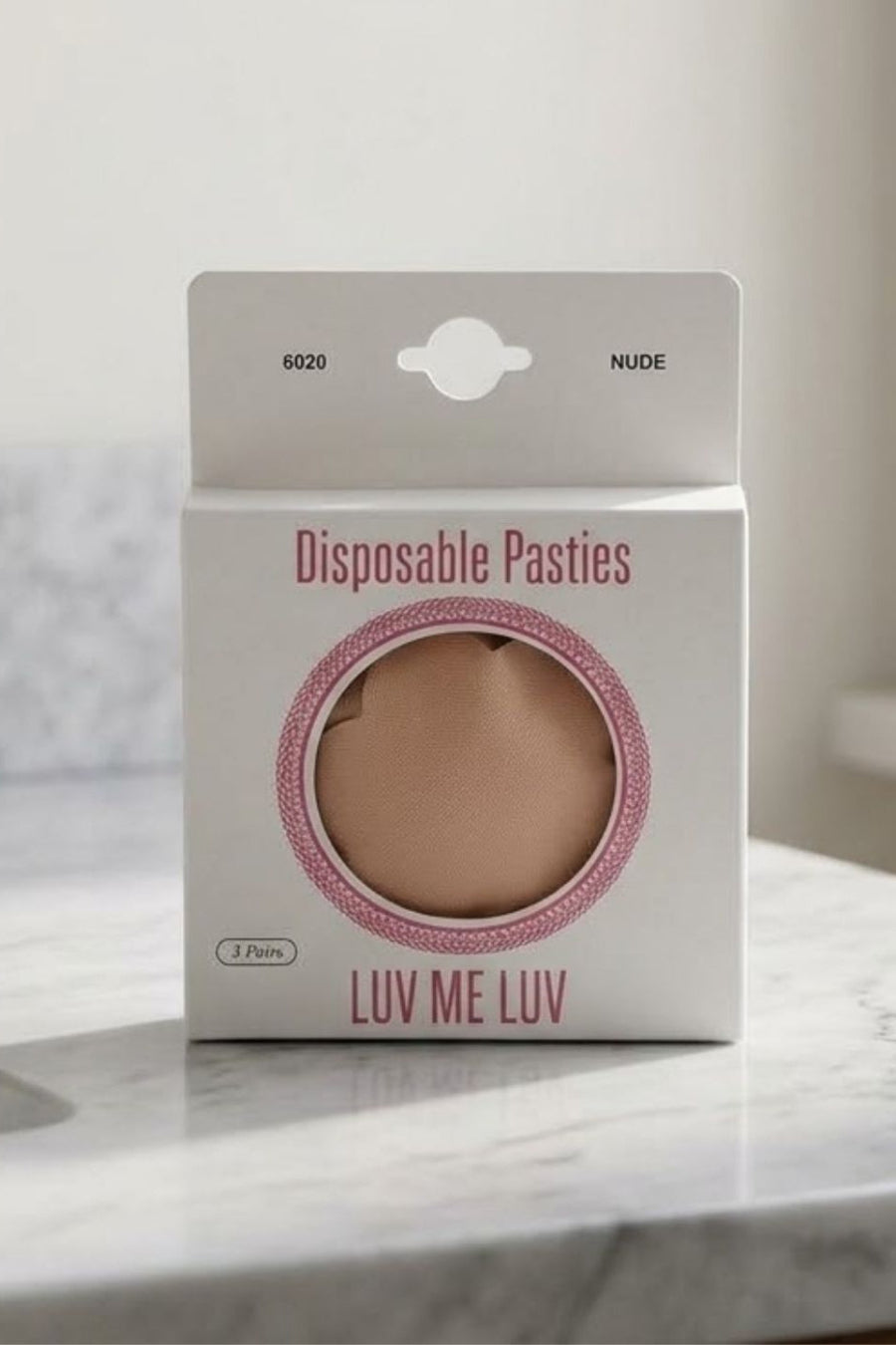 A package of Luv Me Luv disposable pasties in the shade Nude, displayed on a marble countertop with a blurred bathroom background.